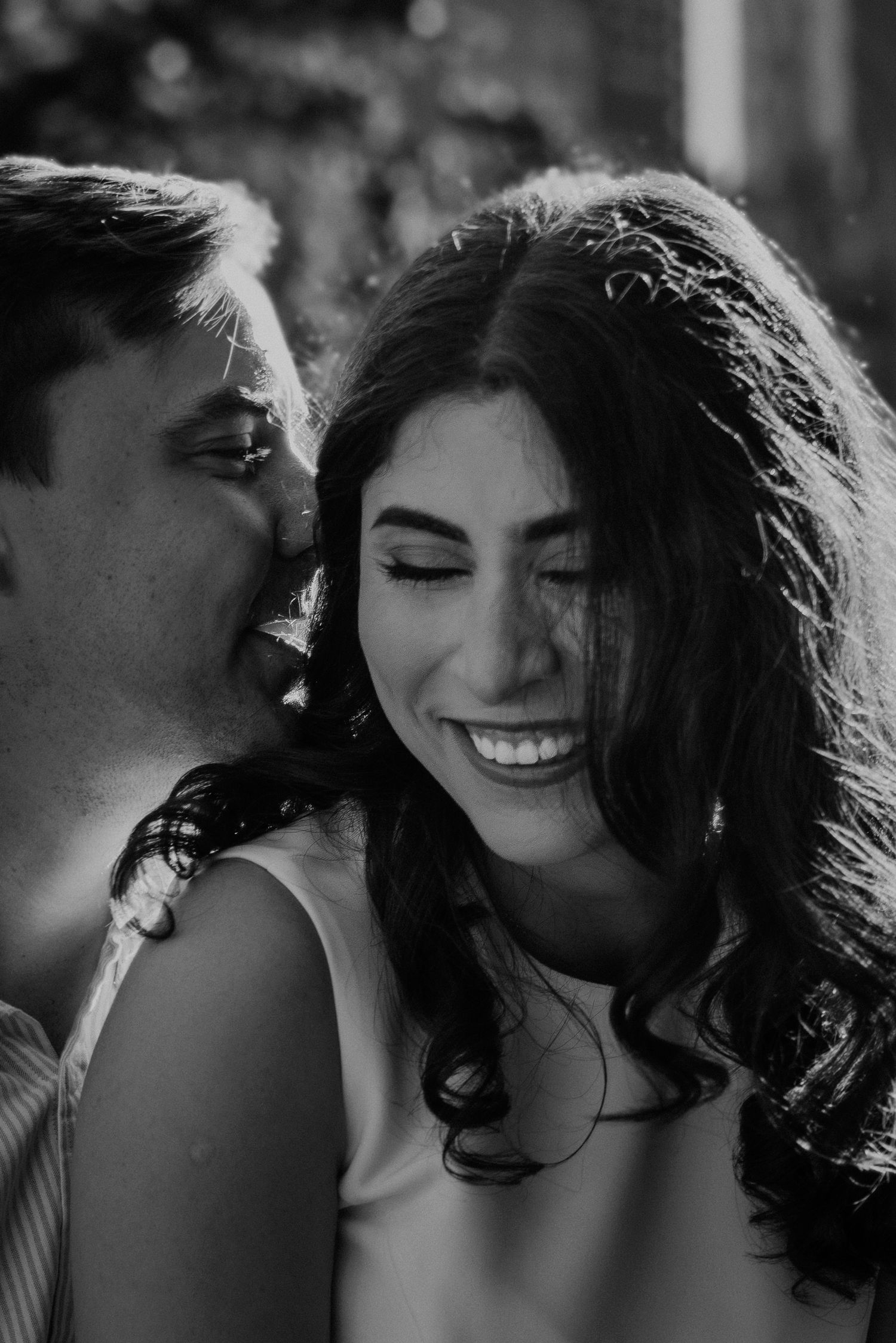 Black and white photo of a smiling woman as a man kisses her cheek during a romantic couples engagement session.