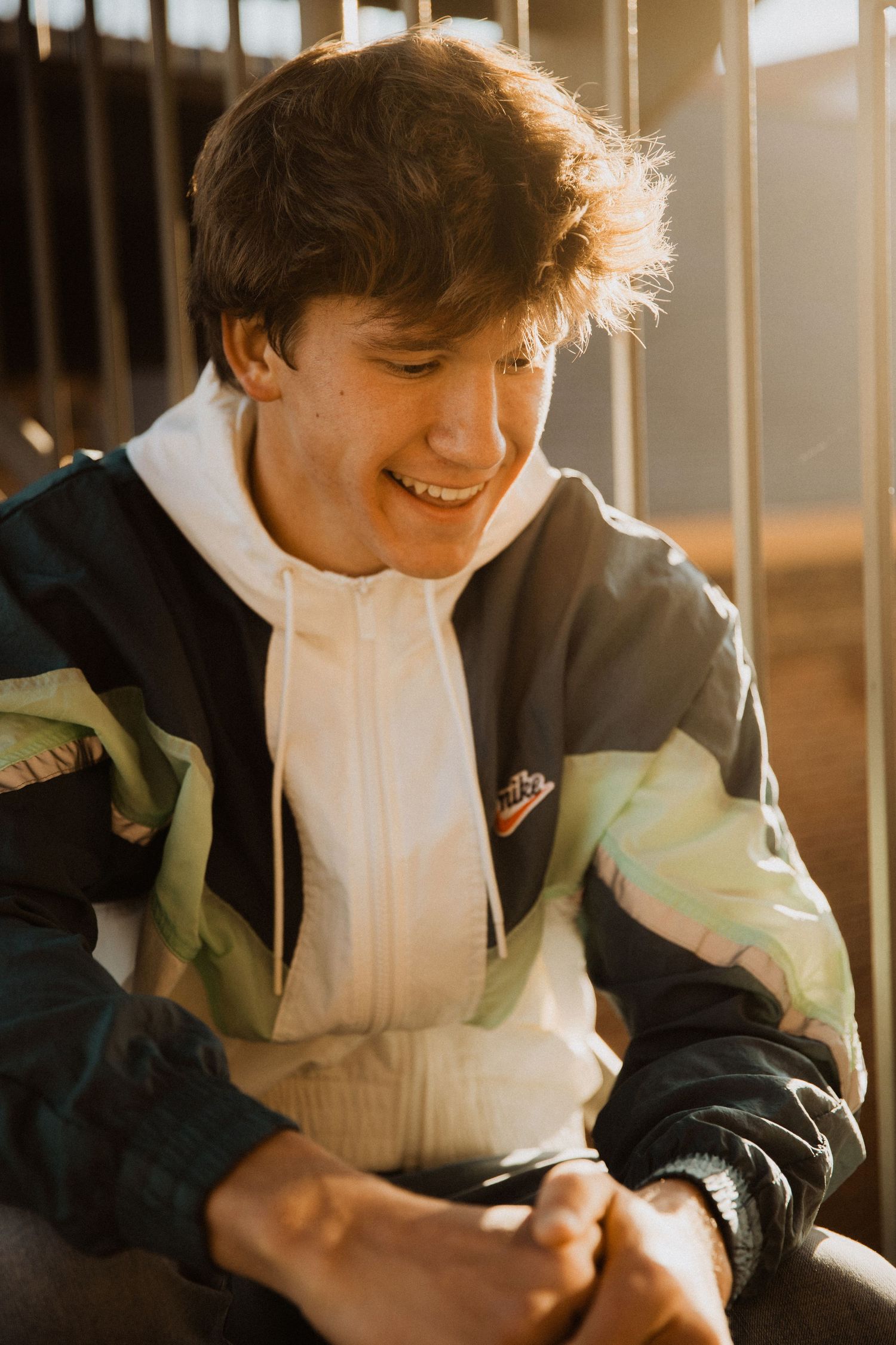 Smiling young man in a retro Nike windbreaker jacket sitting outdoors in warm sunlight.