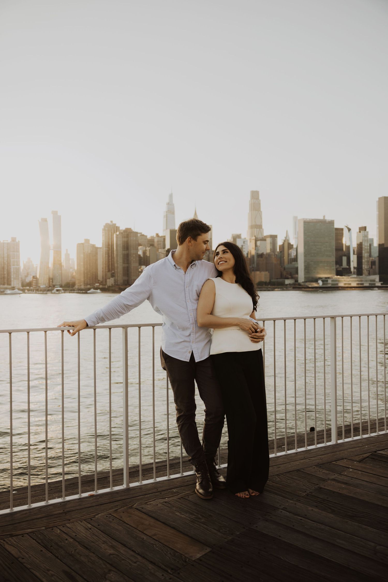 Couple embracing on a waterfront dock with the New York City skyline at golden hour behind them.