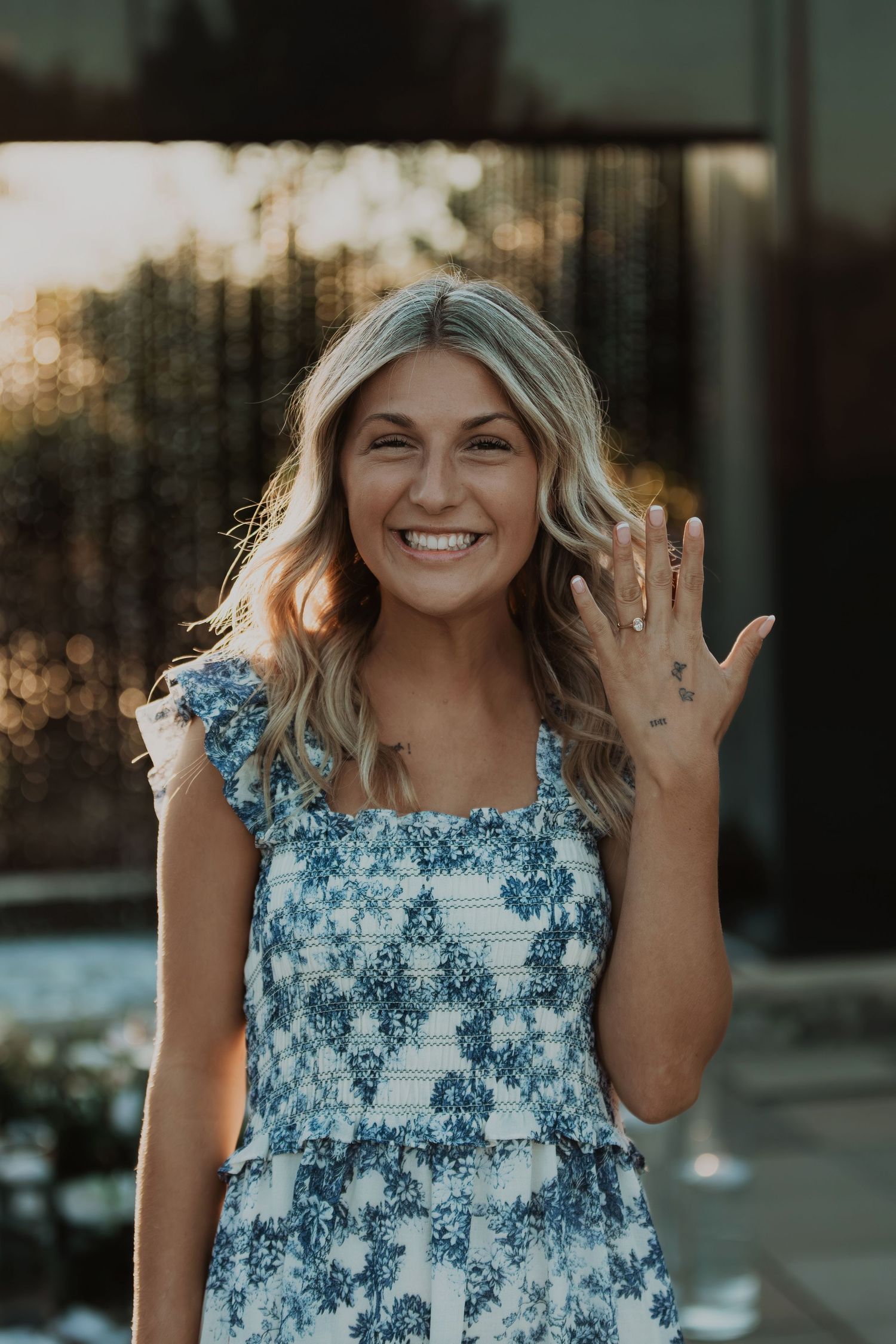 Woman in blue floral dress showing off engagement ring with joyful expression.
