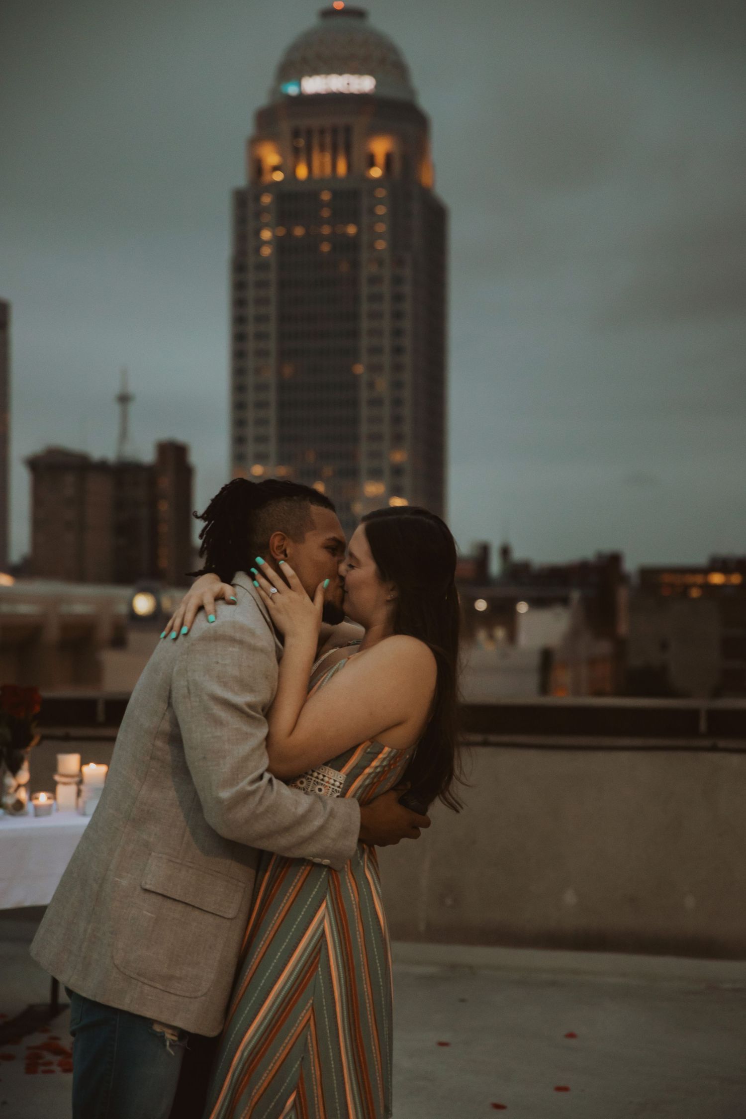 A couple shares a romantic moment on a city rooftop at dusk with illuminated buildings behind them.