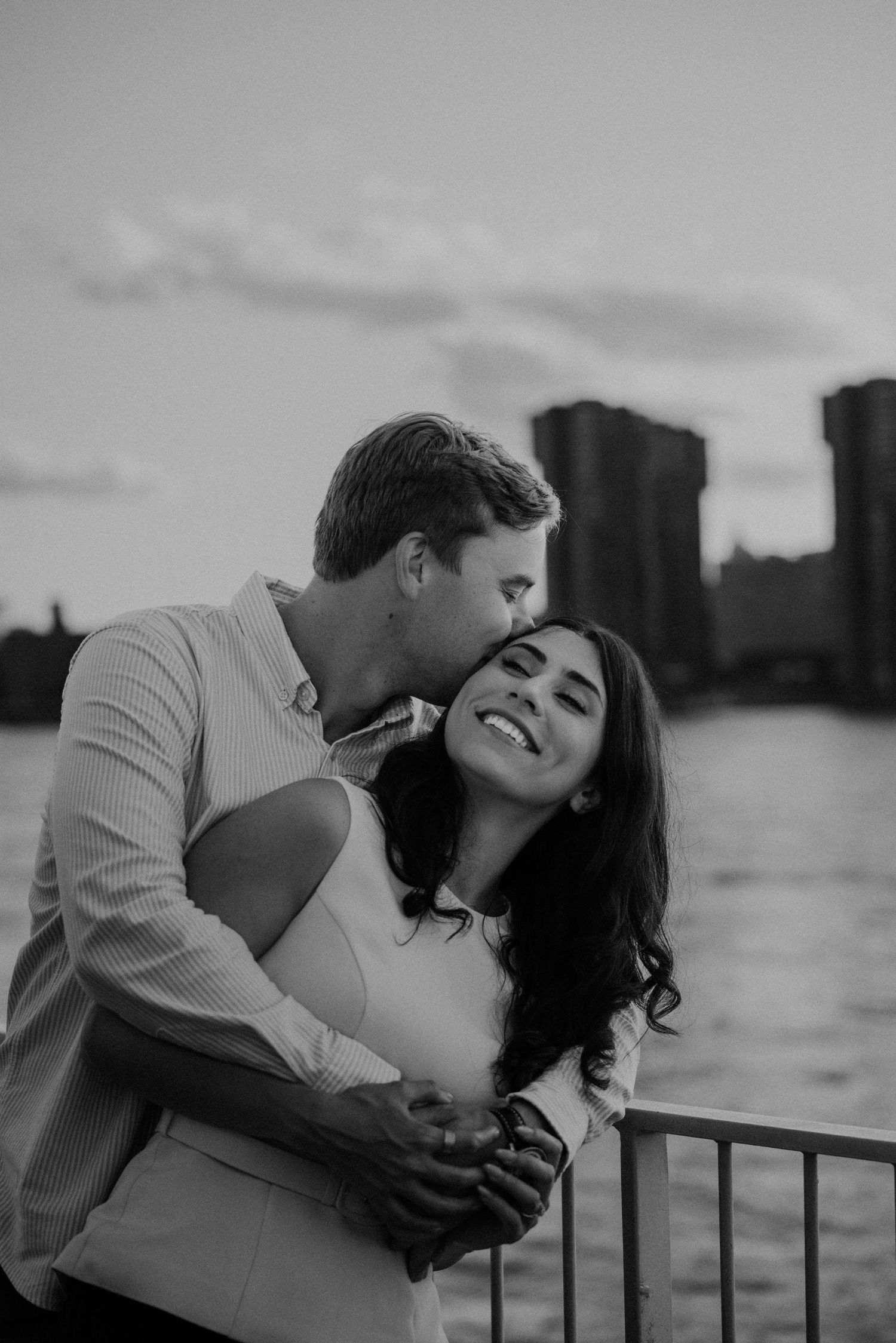Black and white photo of a couple embracing by a waterfront railing at sunset with a city skyline behind them.