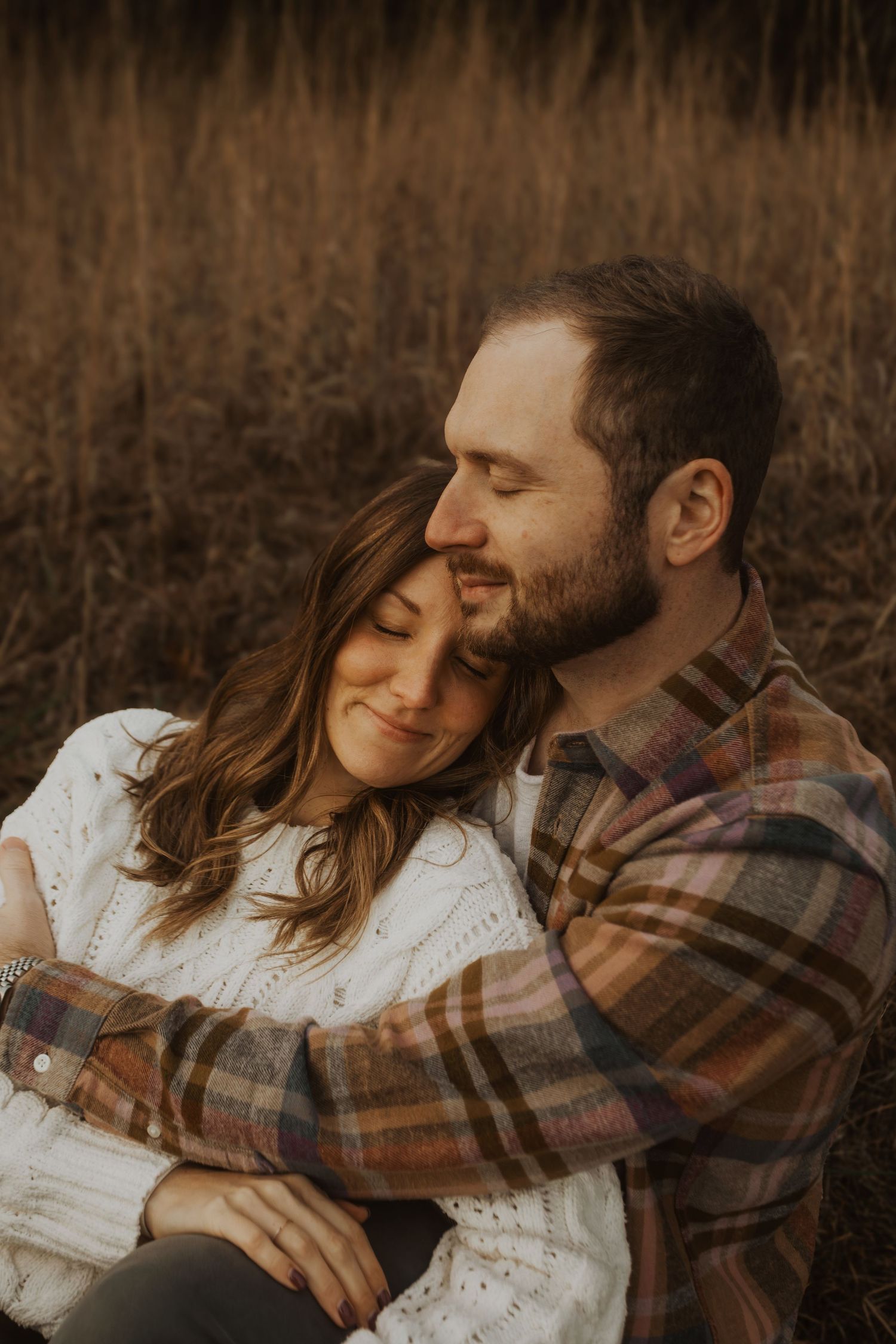 A couple shares a tender embrace in a golden field, the man wrapping his arms around the woman in a white sweater.