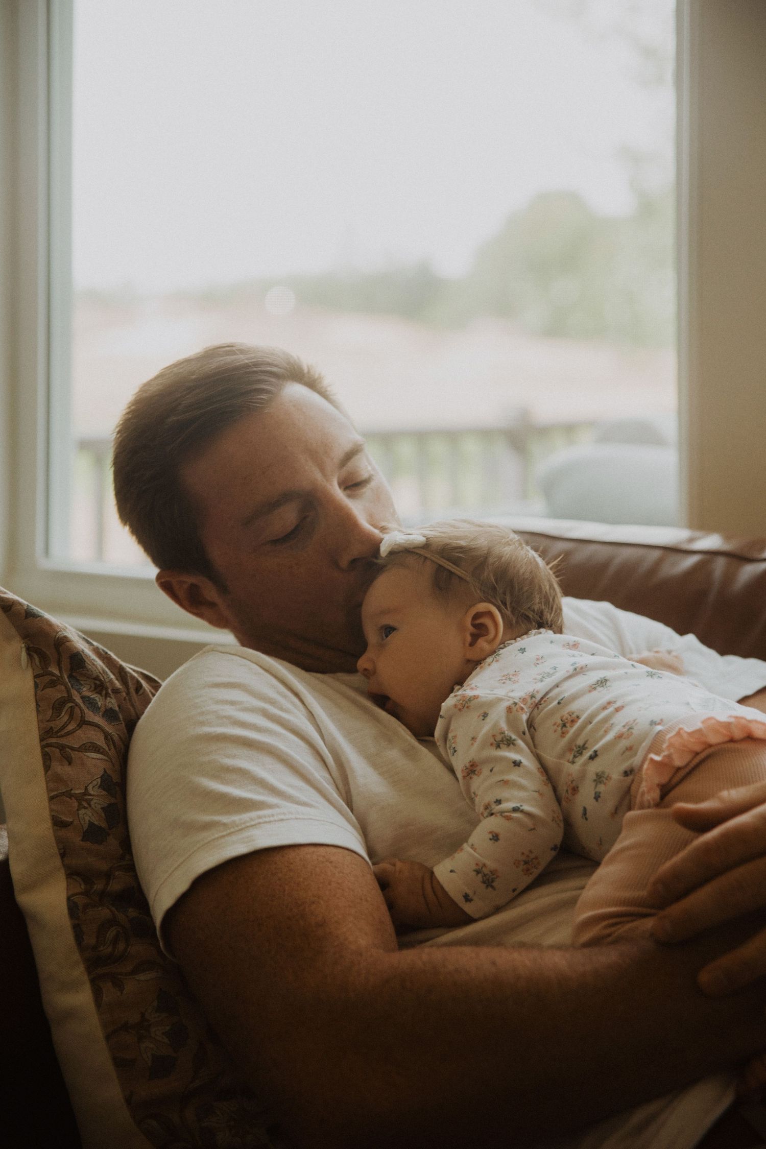 A father tenderly kisses his sleeping baby while resting together on a couch near a bright window.