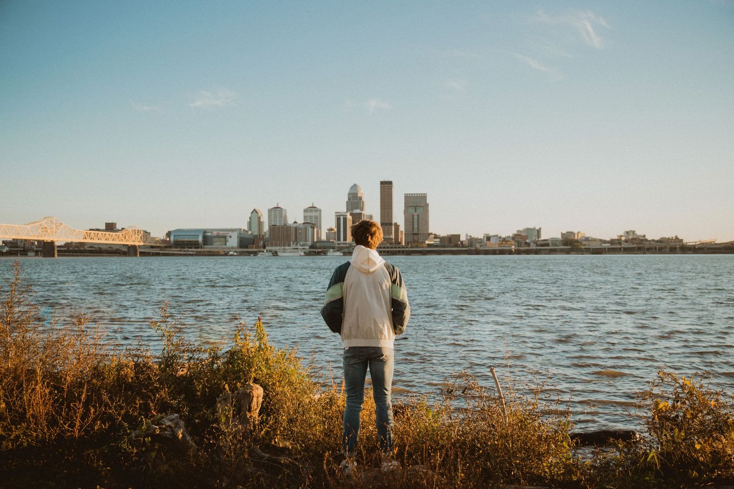 Person standing by a waterfront gazing at a city skyline on a sunny day with lush greenery in the foreground.