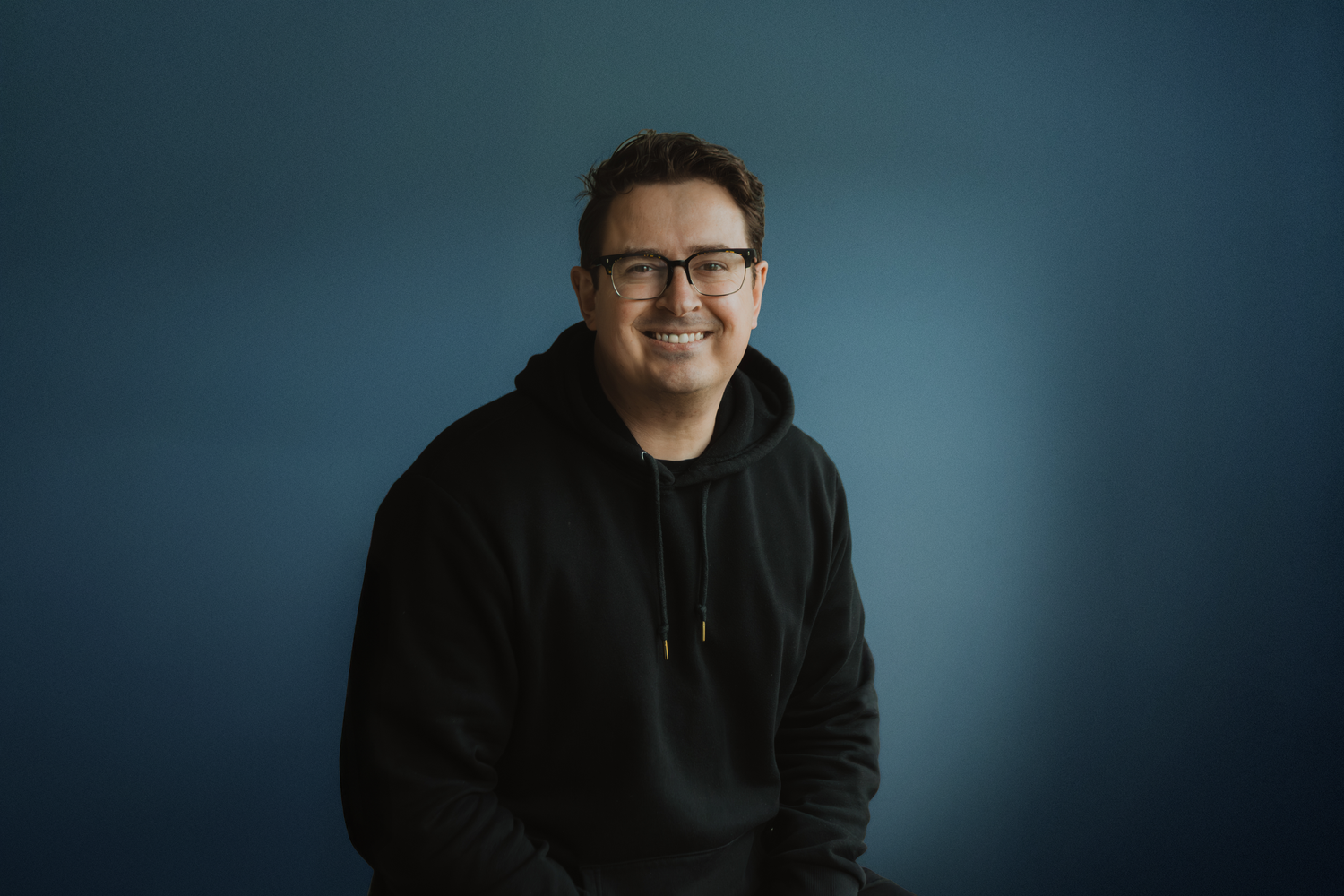 A person wearing glasses and a dark hoodie poses against a blue background in a series of headshots.