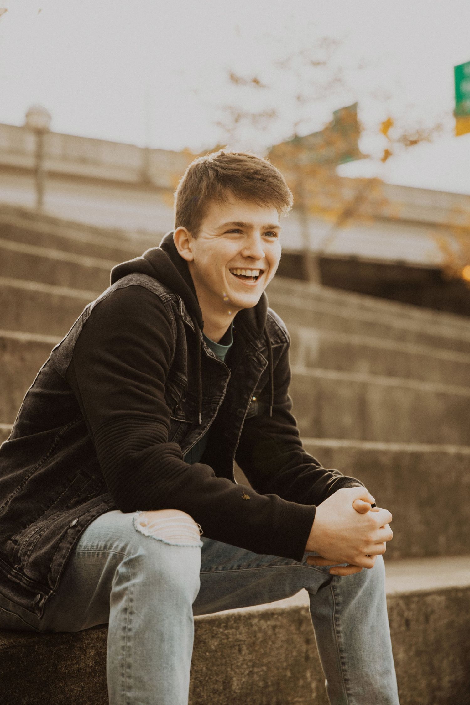 Smiling teenage boy in black hoodie and jeans sitting on stone steps outdoors in warm golden light.