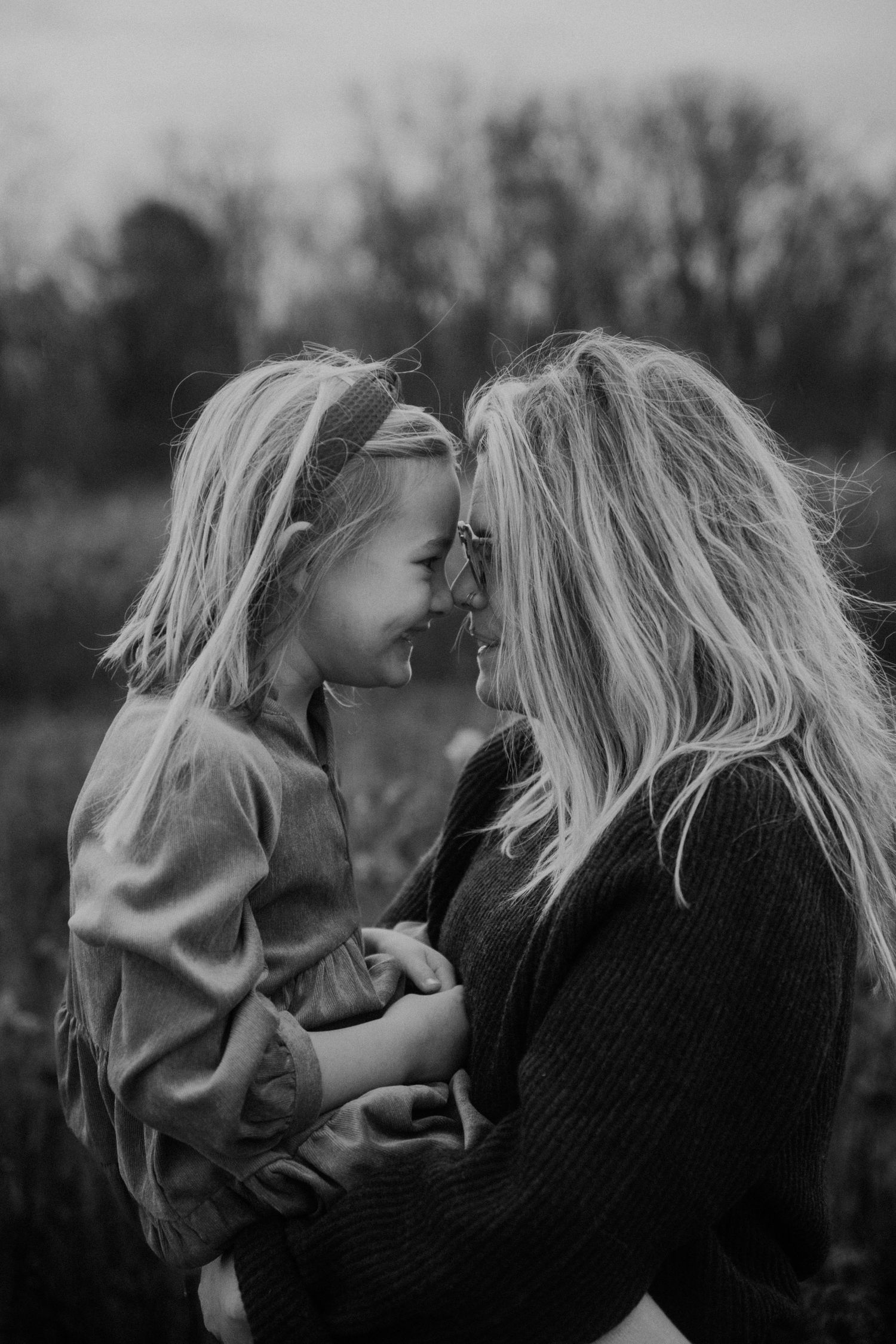 Black and white photo of a mother holding her young blonde daughter, touching foreheads and smiling in an outdoor field.