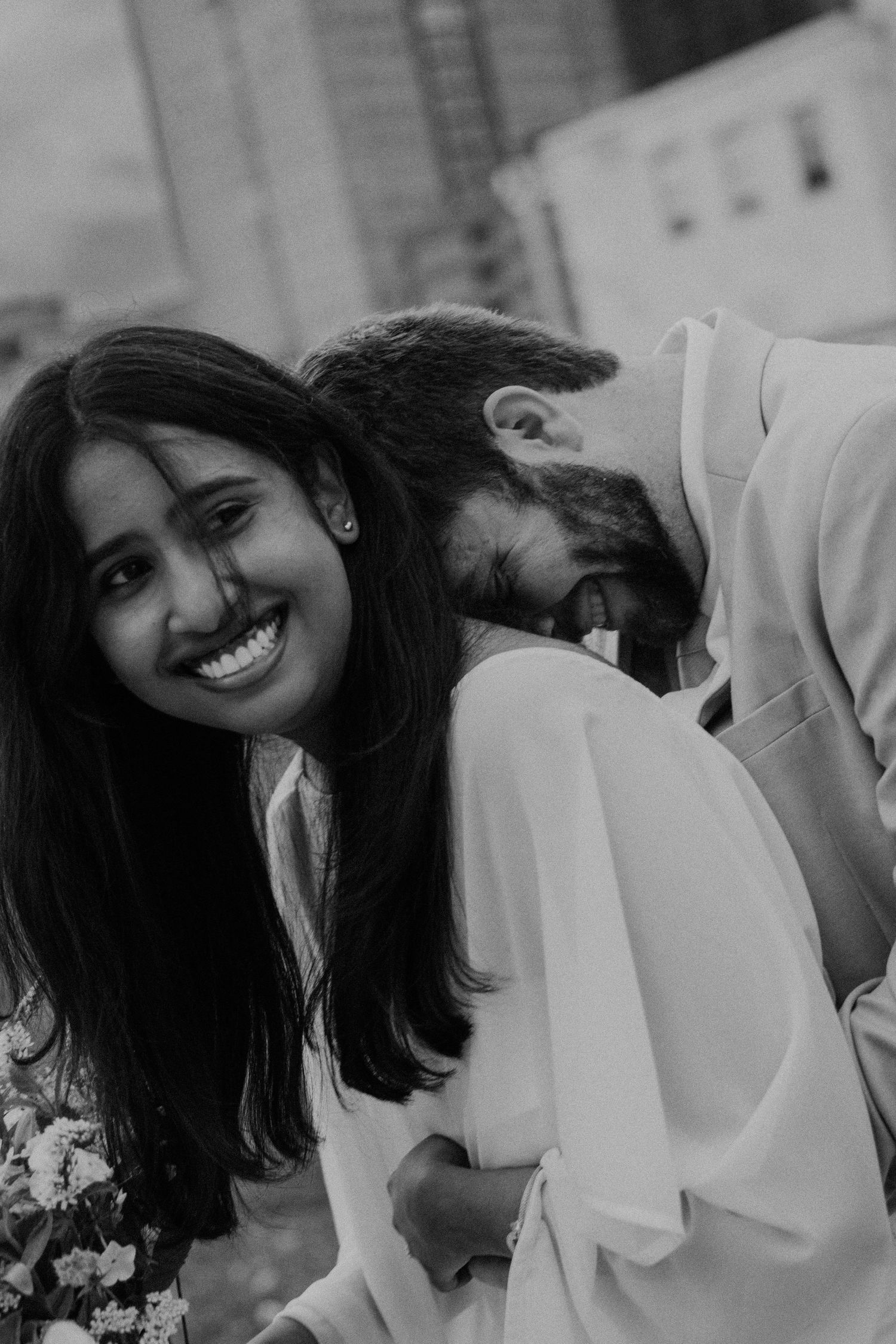 Black and white photo of a smiling South Asian bride leaning against a bearded groom outdoors.