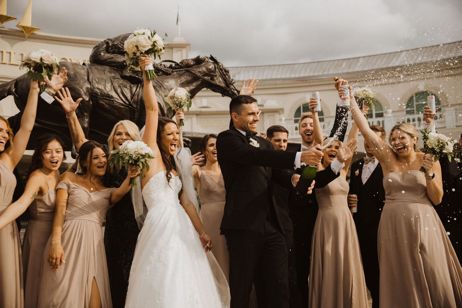 Wedding party celebrates with raised arms and cheers in an elegant indoor venue with arched windows.