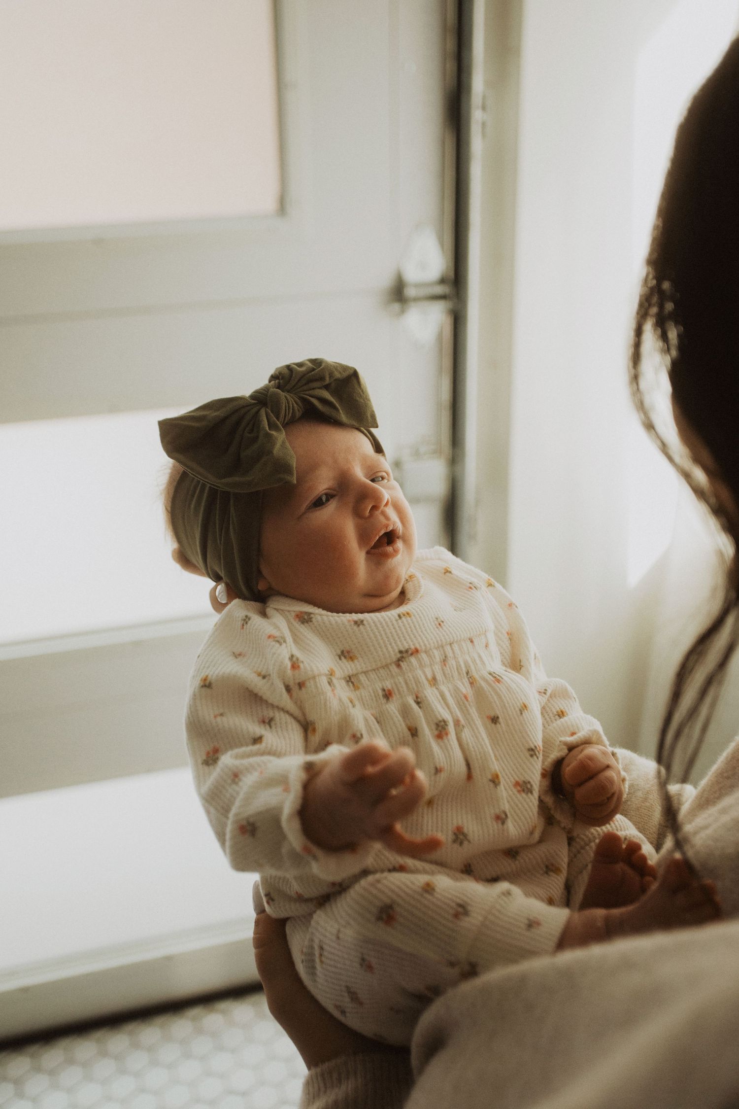 Adorable baby girl in floral romper and olive green bow headband being held near a bright window.