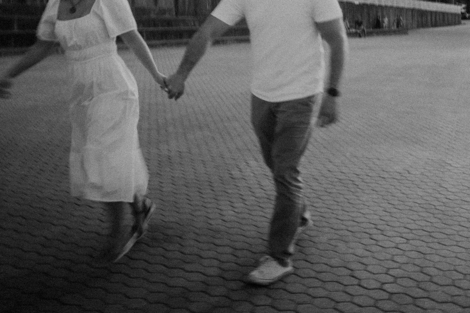 Black and white image of couple holding hands while walking on brick pavement.