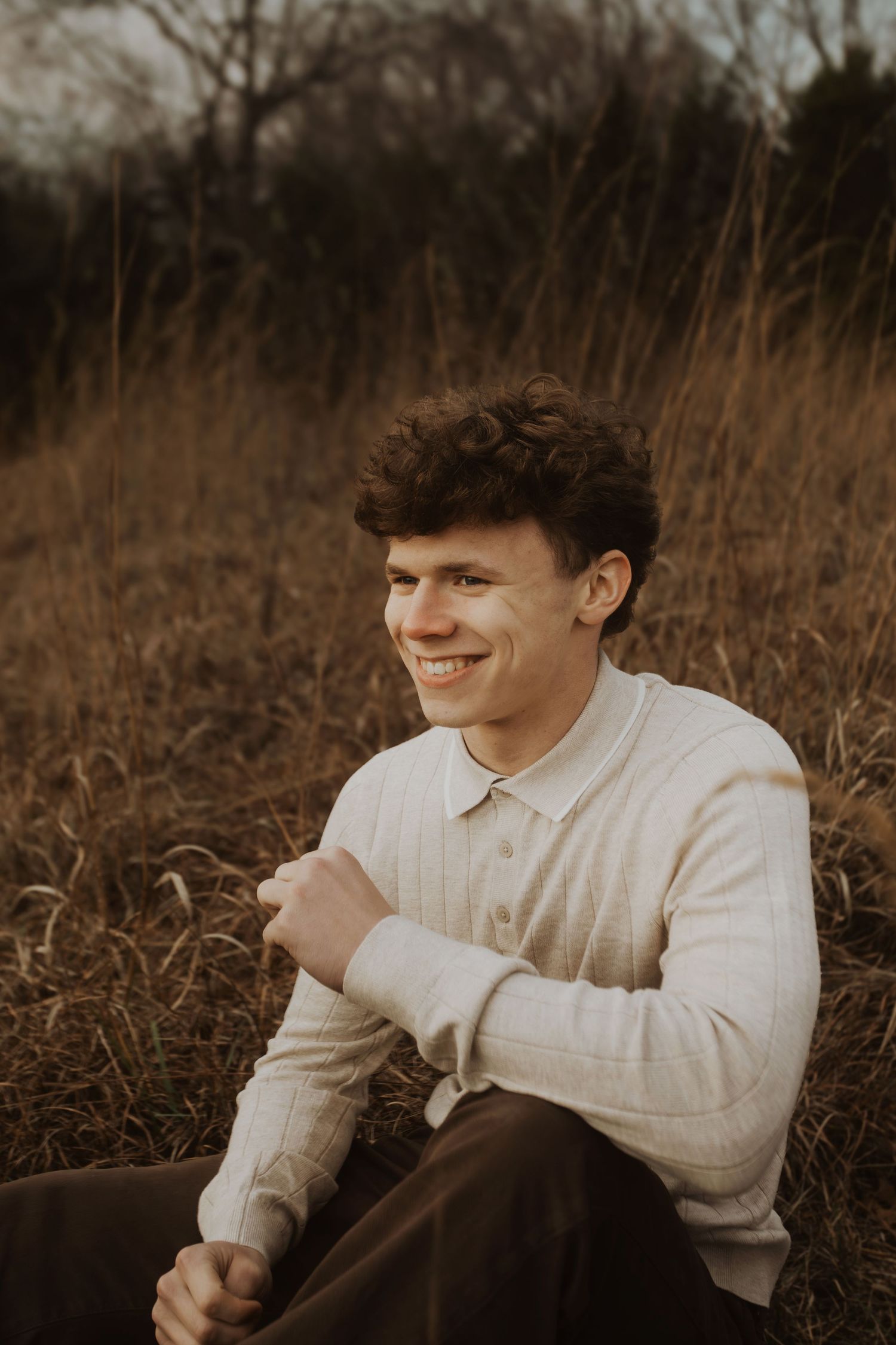 Smiling young man with curly hair sitting in a field of tall dry grass wearing a cream ribbed polo sweater.