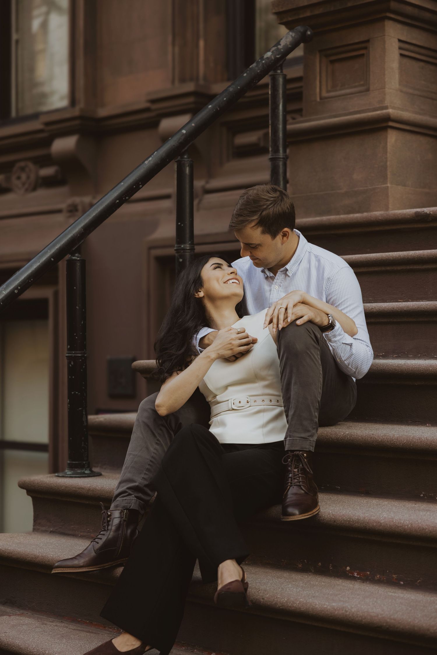 Couple sitting on brownstone steps in NYC, woman leaning back smiling up at man in blue shirt in a romantic pose.