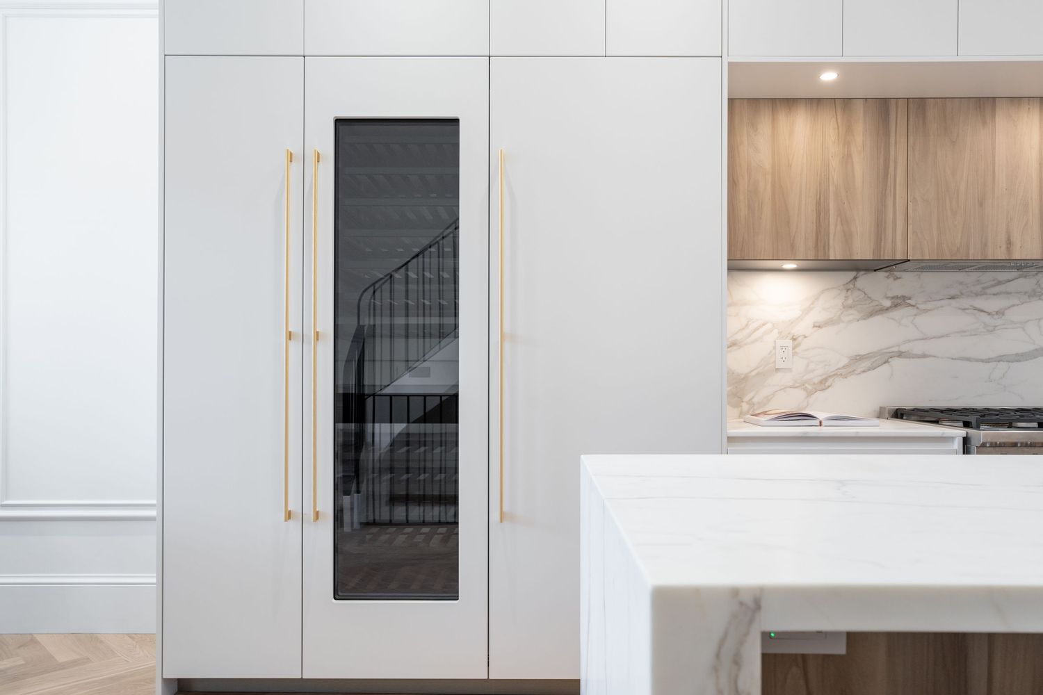 Contemporary white kitchen with gold hardware, marble island, wood upper cabinets, and integrated appliances.