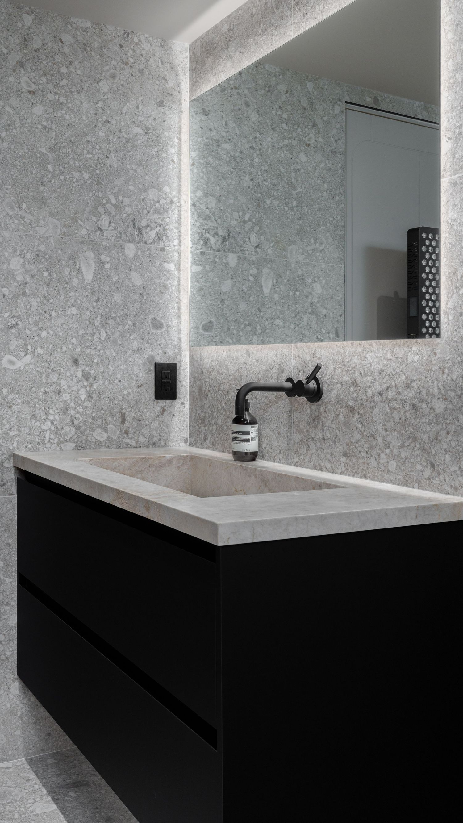 Sleek black floating vanity with marble sink, backlit mirror, and terrazzo wall tiles.