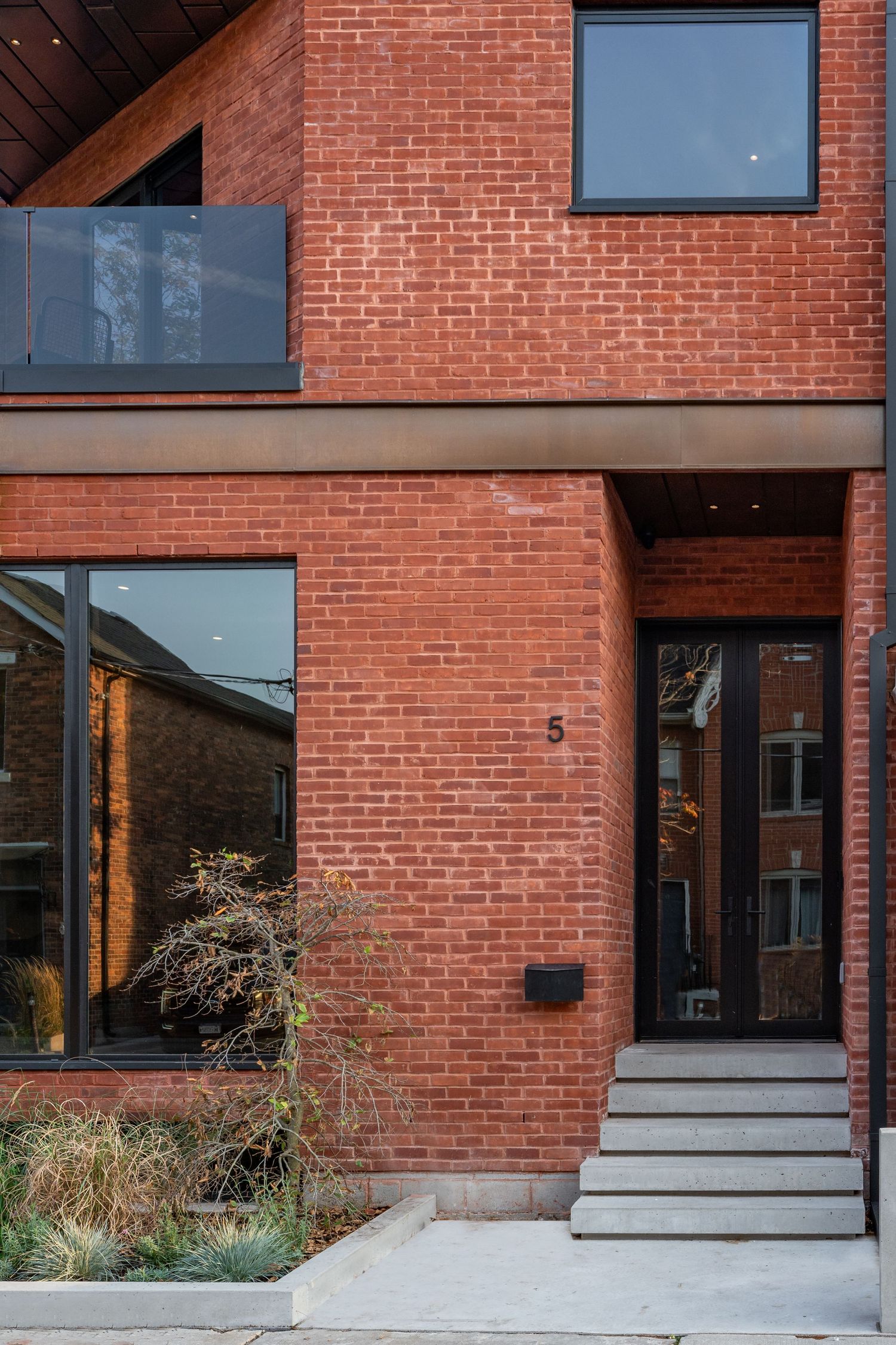 Modern red brick townhouse exterior with large black-framed windows, concrete steps, and a wood front door numbered 5.