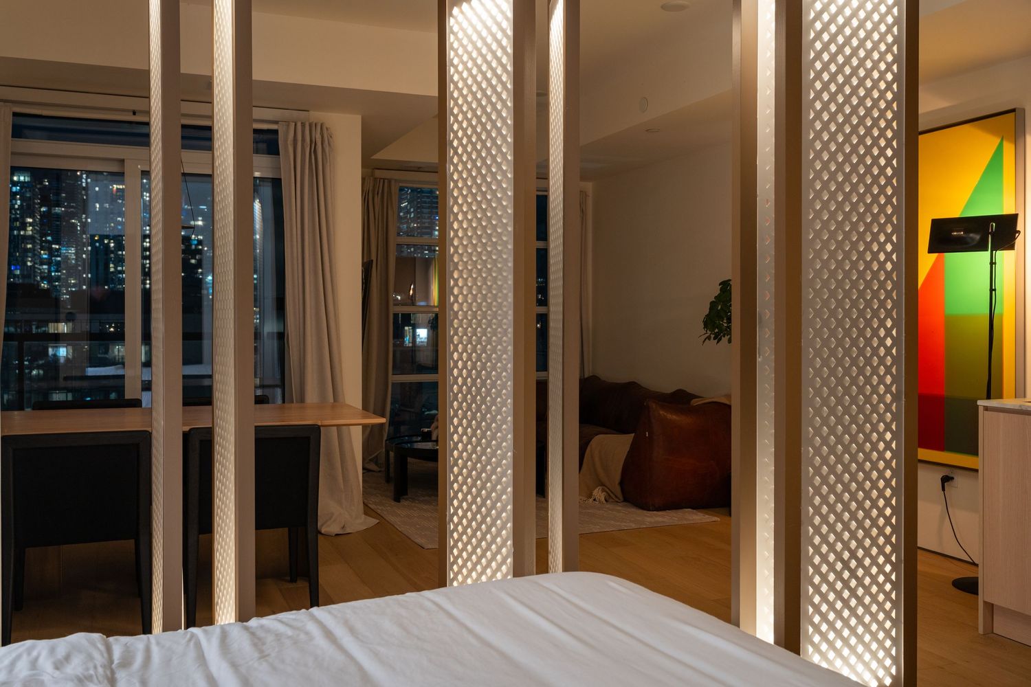 Contemporary bedroom with illuminated lattice room dividers, city night view, hardwood floors, and colorful accent wall.