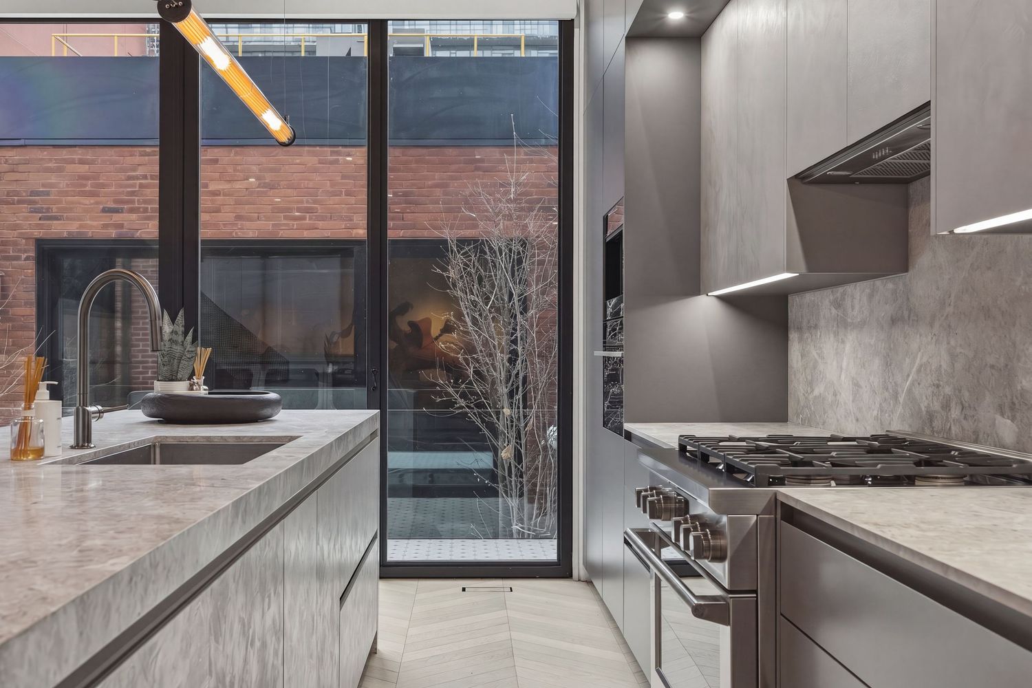 Modern kitchen with marble countertops, gray cabinets, gas range, and large windows overlooking a brick courtyard.