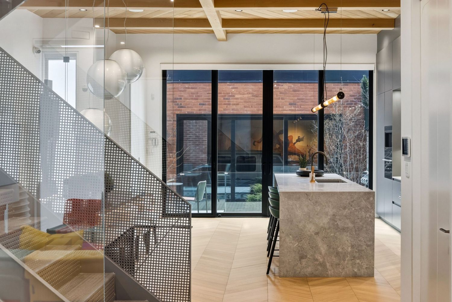 Modern kitchen with marble island, herringbone floors, glass staircase, and floor-to-ceiling windows overlooking brick courtyard.