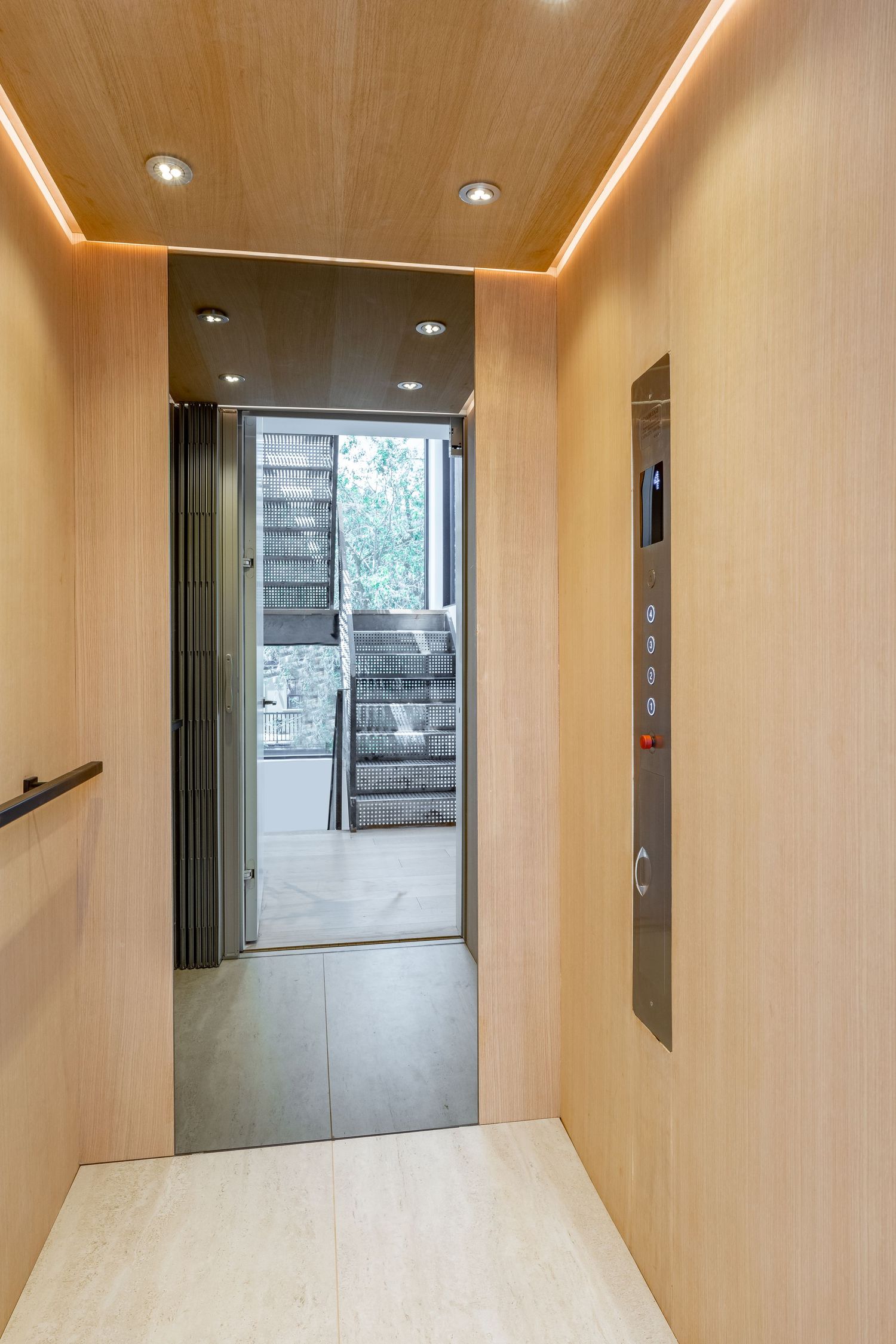 Modern elevator interior with light wood paneling, recessed lighting, and open doors revealing a staircase beyond.