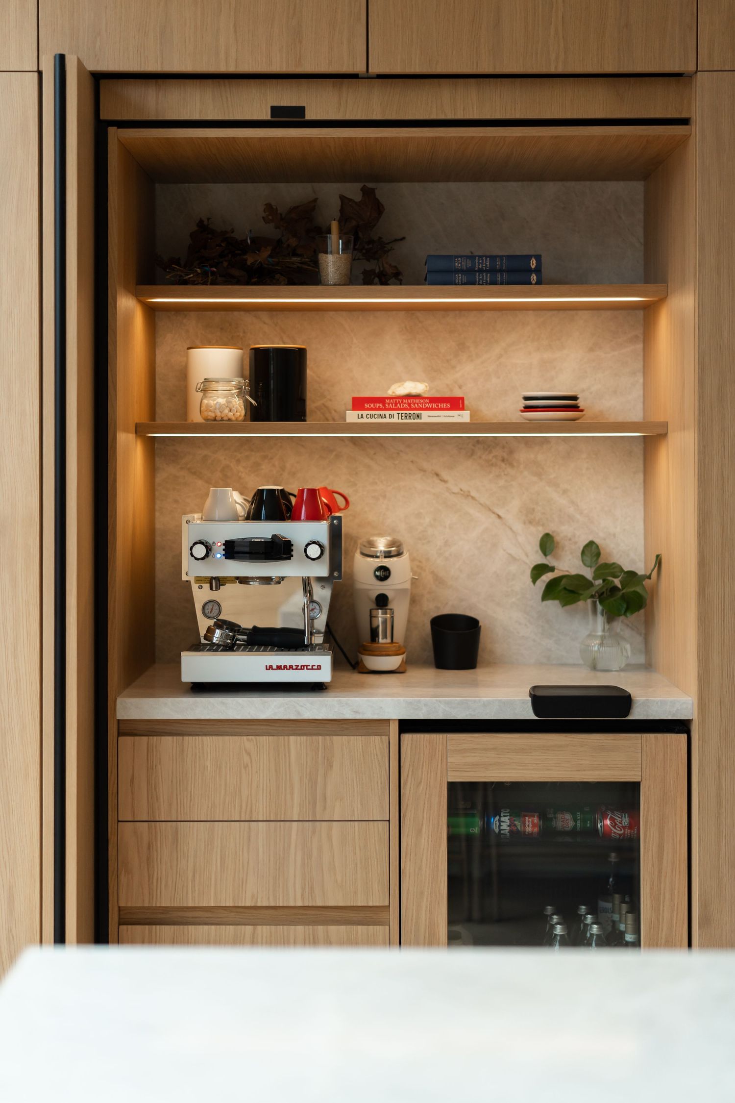 Built-in wooden coffee station with marble shelving, espresso machine, wine fridge, and warm under-cabinet lighting.
