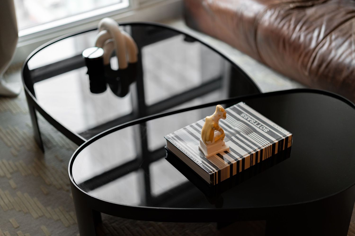 Black oval glass coffee table with decorative sculptures and striped book on dark floor.