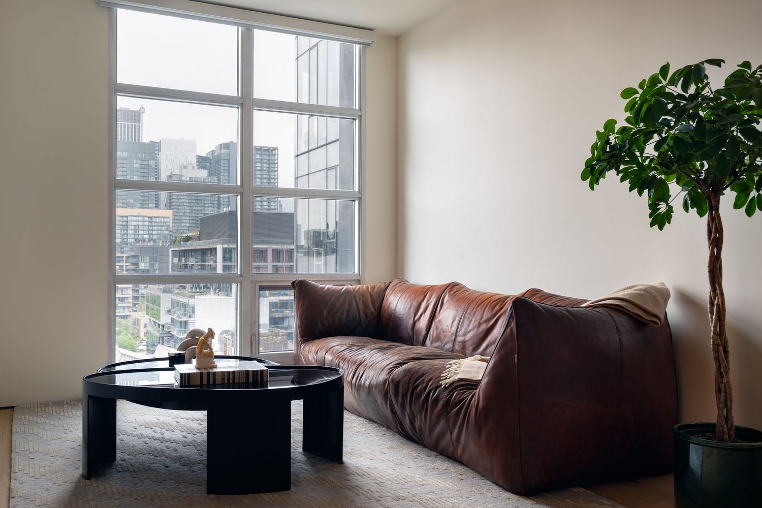 Bright corner living room with large windows, brown leather sofa, black coffee table, and city views.