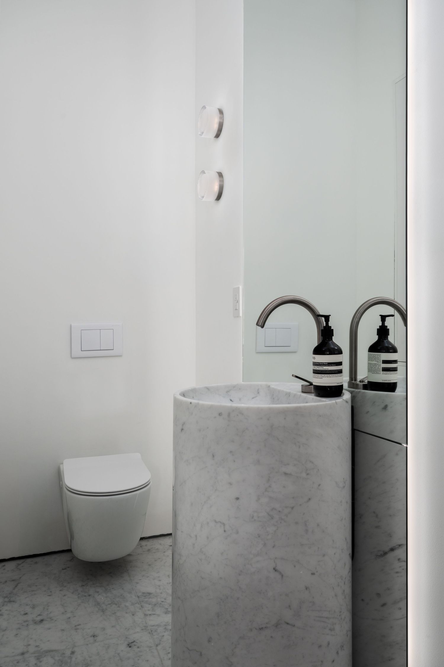 Minimalist bathroom with cylindrical marble pedestal sink, wall-mounted faucet, and large mirror.