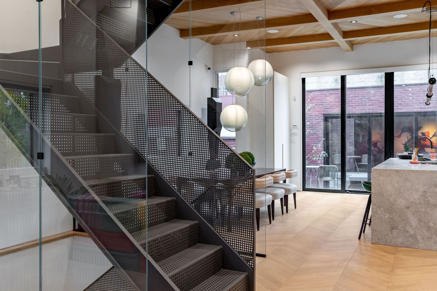 Modern open-plan interior featuring glass staircase, herringbone wood floors, pendant lights, and floor-to-ceiling windows.
