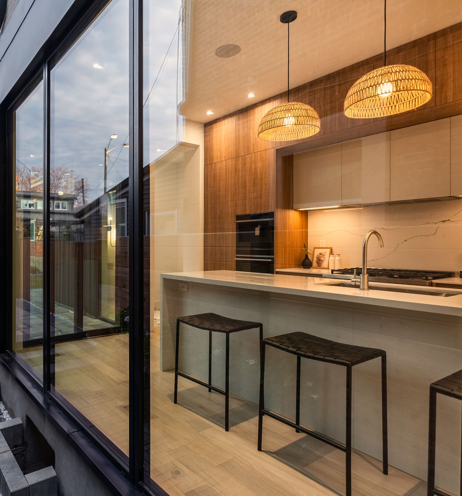 Modern kitchen with pendant lights, marble island, bar stools, and floor-to-ceiling glass walls overlooking an outdoor area.