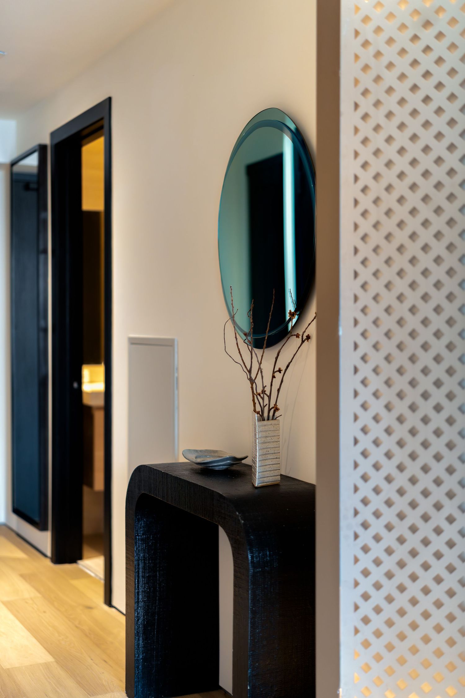 Modern hallway with oval teal mirror, black console table, decorative lattice wall panel, and wood floors.