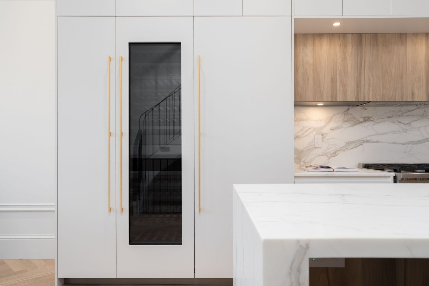 Modern white kitchen with gold handles, marble backsplash, and sleek cabinetry featuring a dark glass panel accent.