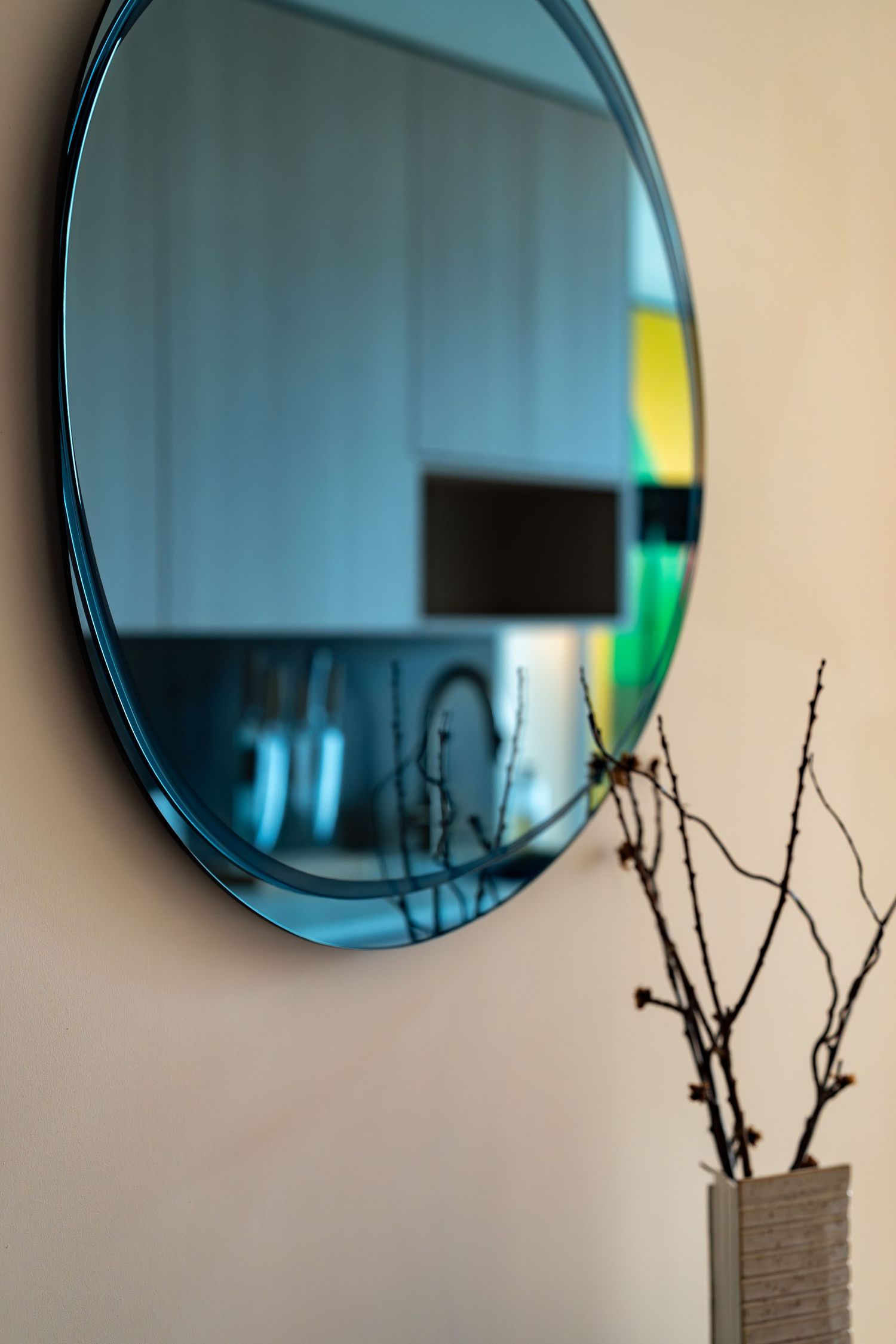 Round blue-tinted mirror on beige wall beside a vase with bare branches.