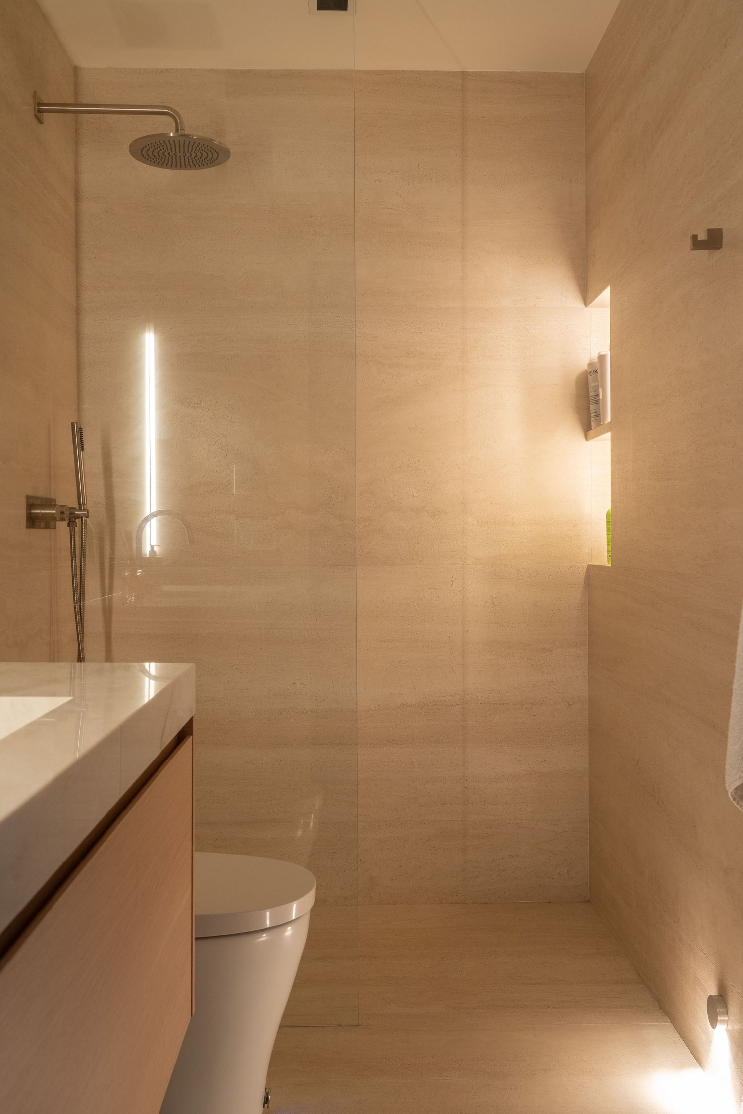 Elegant beige-tiled bathroom with rain shower, glass partition, and warm accent lighting.