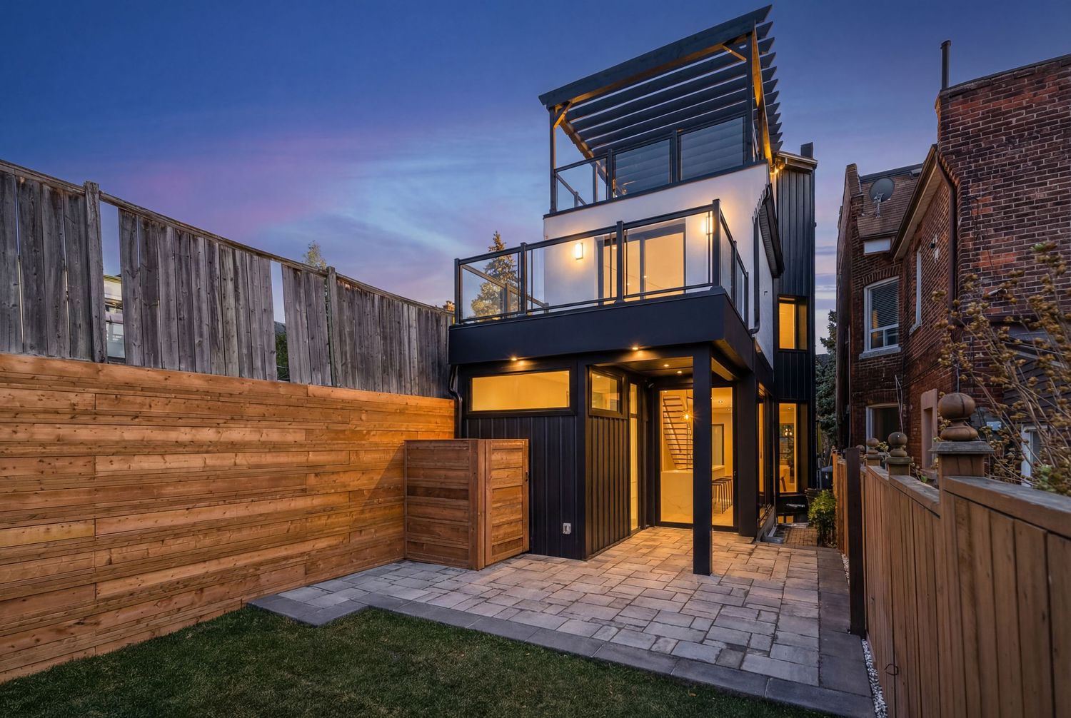 Modern two-story home with rooftop deck, black exterior, glass doors, and patio at dusk with purple sky.