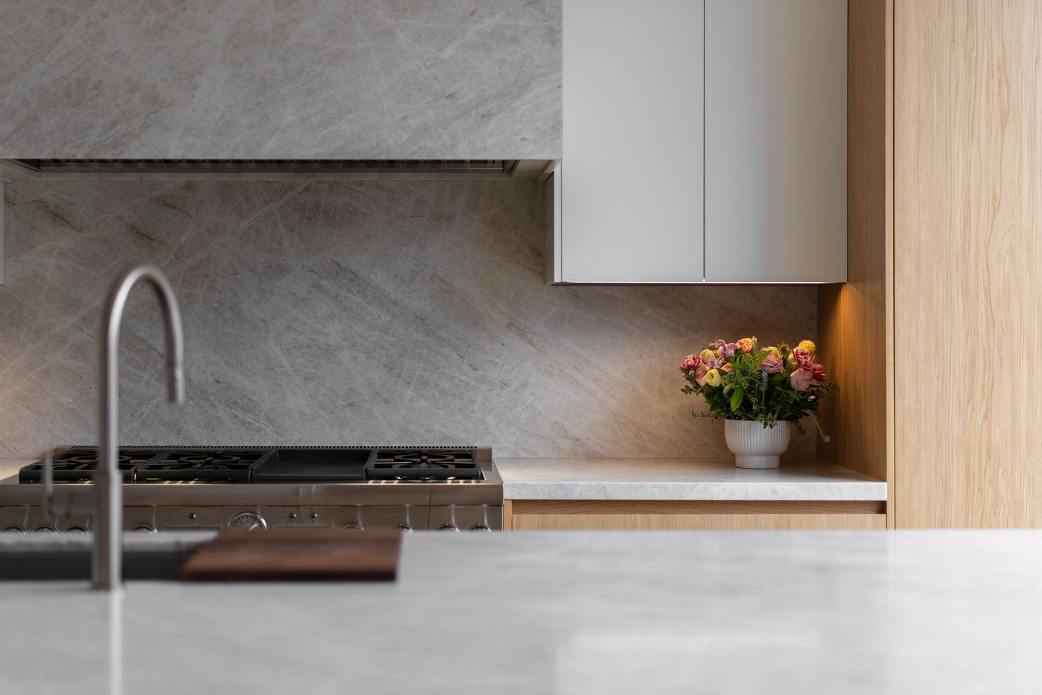 Close-up of modern kitchen with grey marble backsplash, gas cooktop, fresh flowers, and warm wooden cabinet accents.