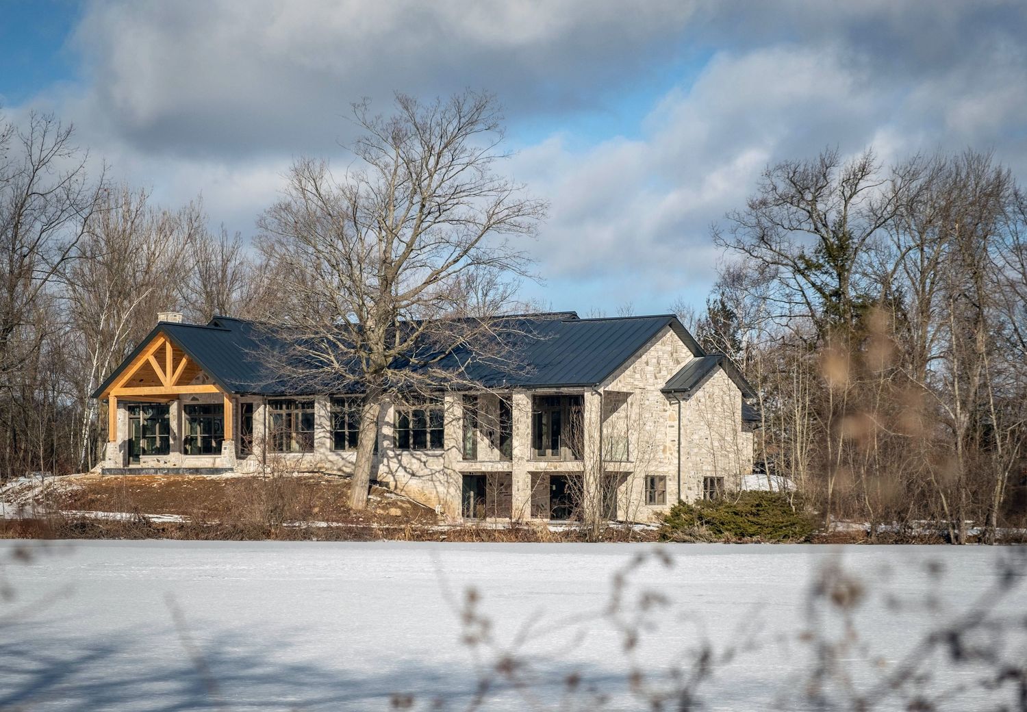 Stunning stone and timber luxury home exterior surrounded by snow-covered landscape and bare winter trees.