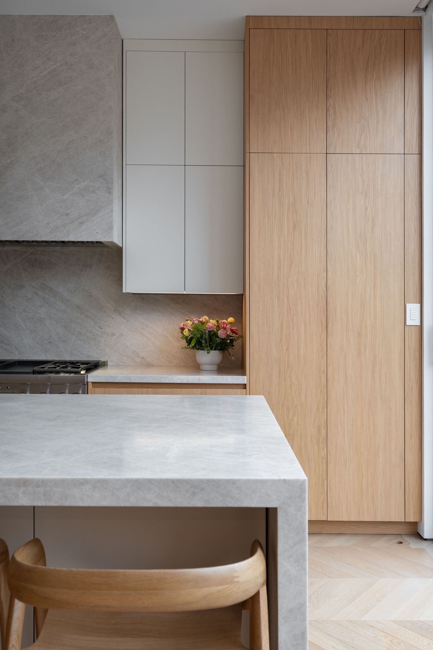 Modern kitchen detail showing grey marble island, white upper cabinets, natural oak pantry, and herringbone wood floor.