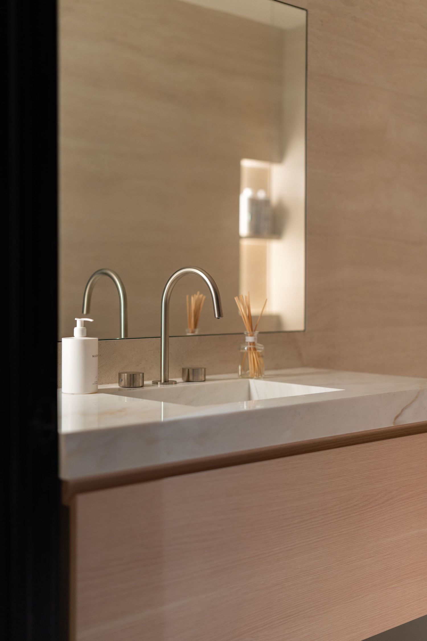 Modern bathroom vanity with marble countertop, brushed nickel faucets, reed diffuser, and large mirror on beige stone wall.