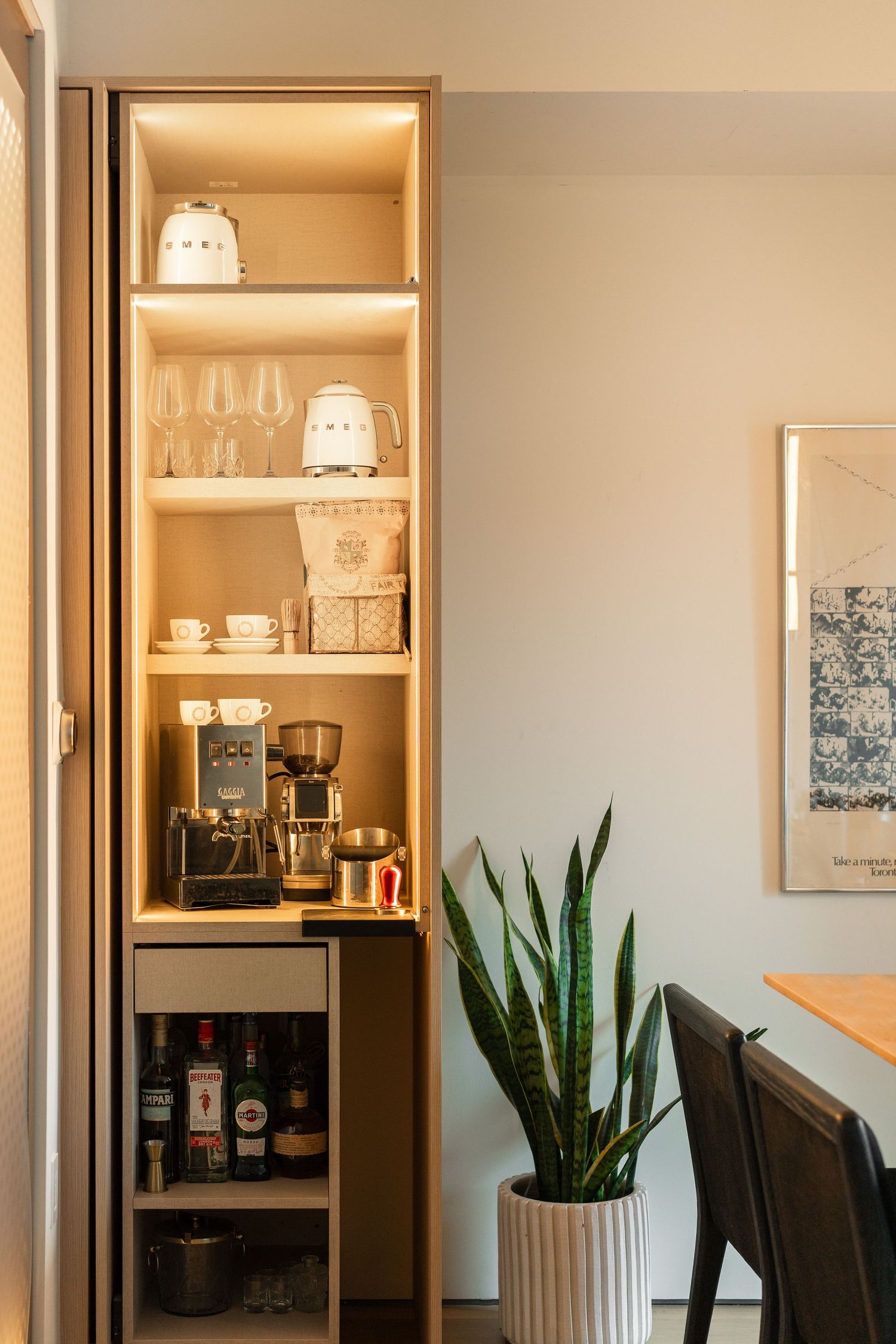 Built-in wooden bar cabinet with espresso machine, glassware, and bottles beside a snake plant.