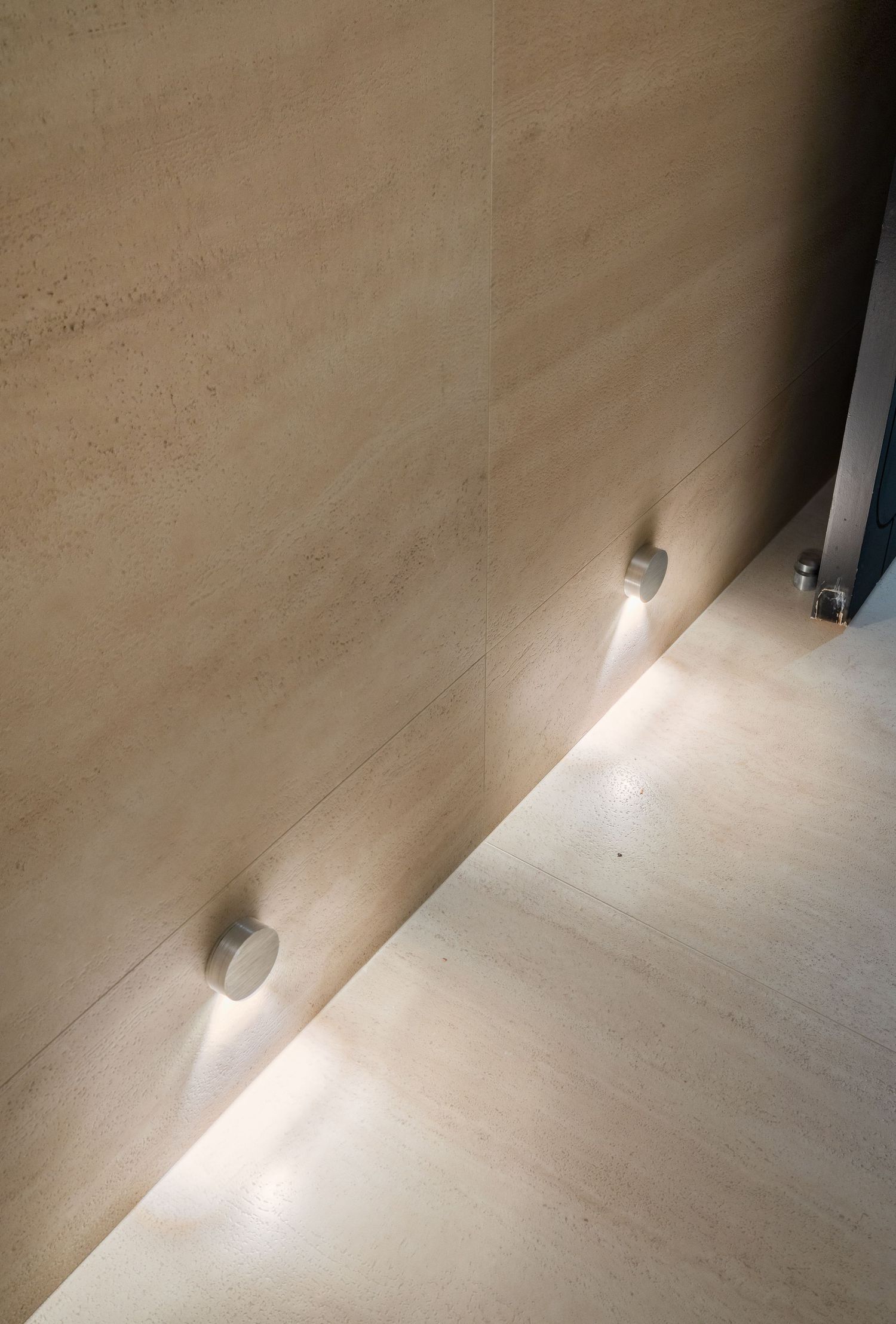 Minimalist beige stone wall with two small round wall-mounted accent lights casting downward glow.