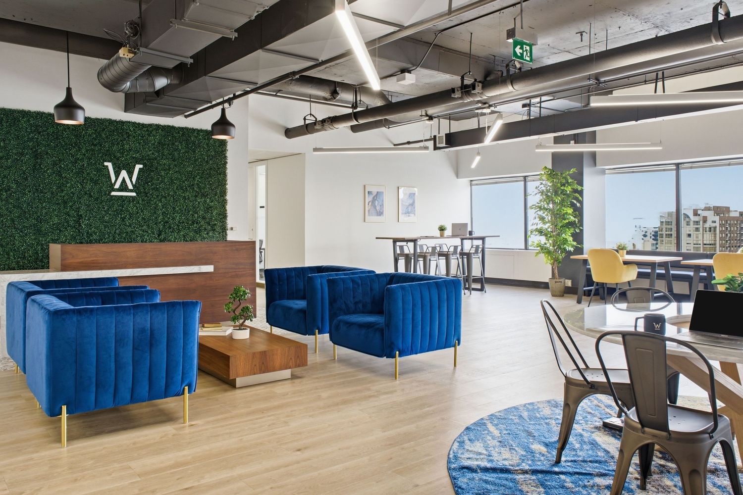 Modern coworking lounge with blue velvet chairs, green moss wall, and open workspace with large windows.