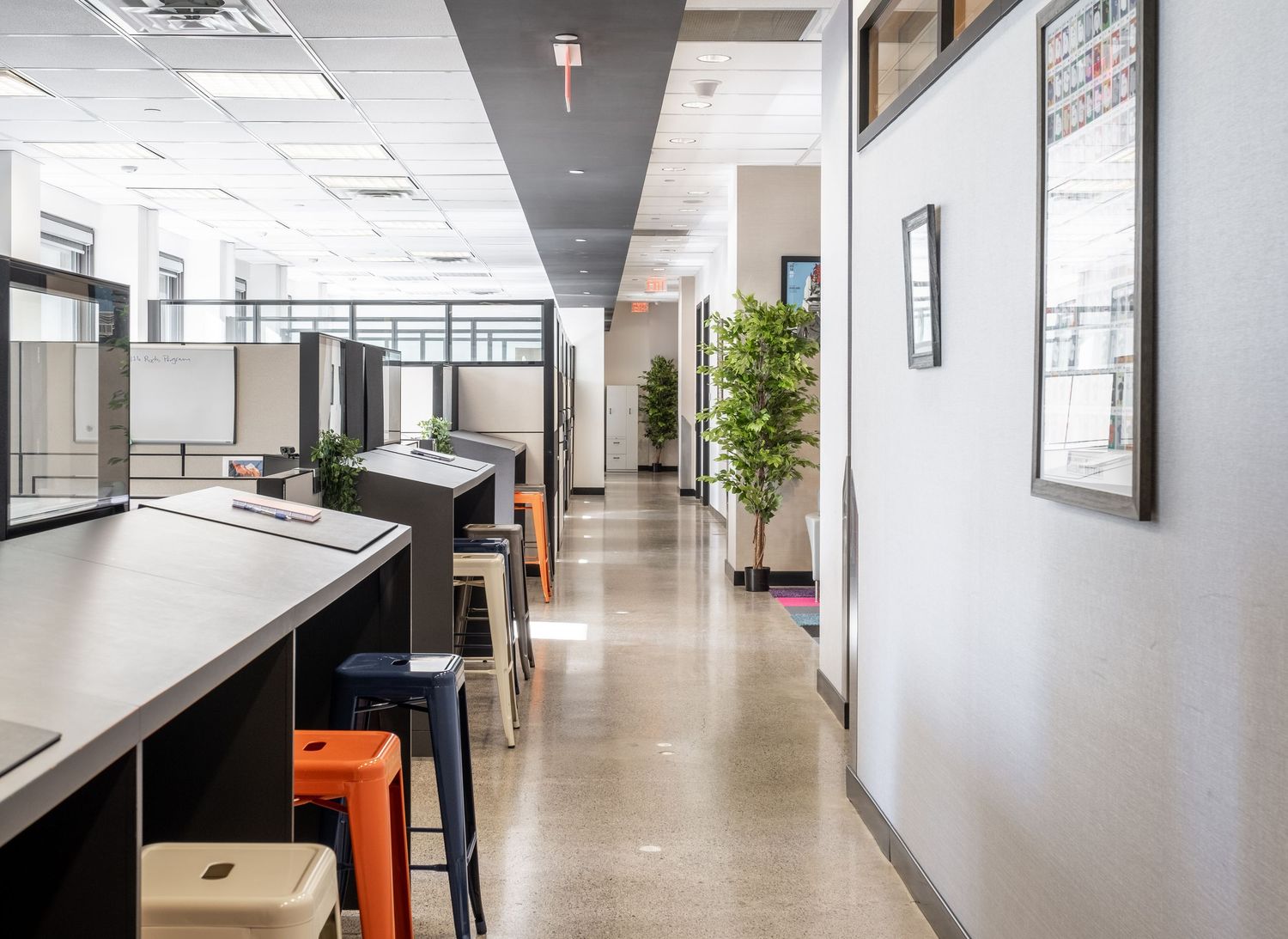 Modern open-plan office with cubicles, colorful stools, polished floors, and green plants along a bright corridor.