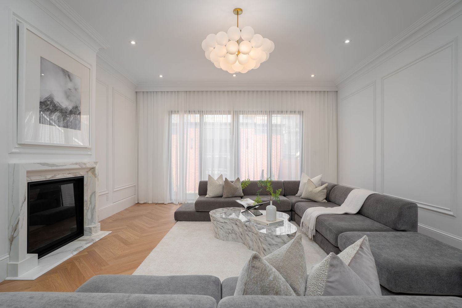 Elegant living room with gray sectional sofa, marble fireplace, herringbone floors, and a glass bubble chandelier.