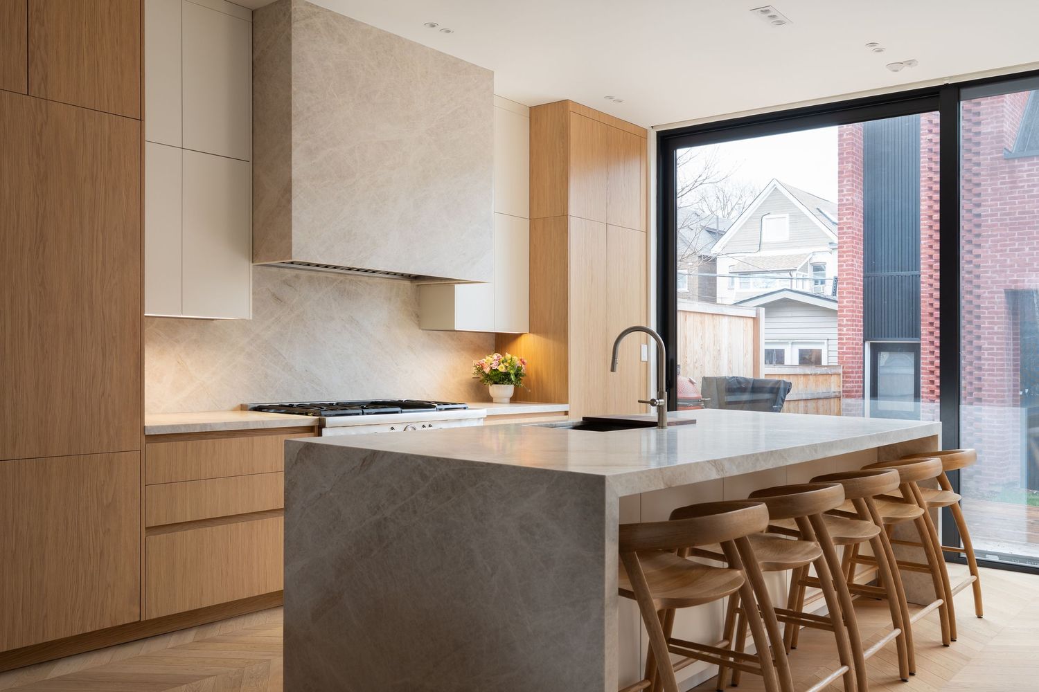 Modern kitchen with concrete island, wood cabinets, bar stools, and large window overlooking neighborhood.