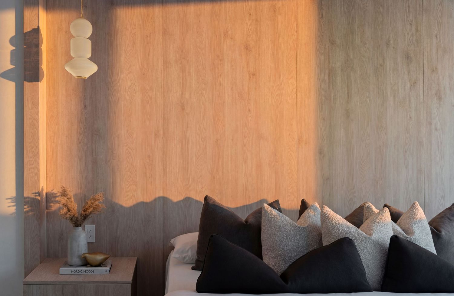 Cozy bedroom with wood panel wall, dark throw pillows on bed, warm ambient lighting, and a decorative pendant light.