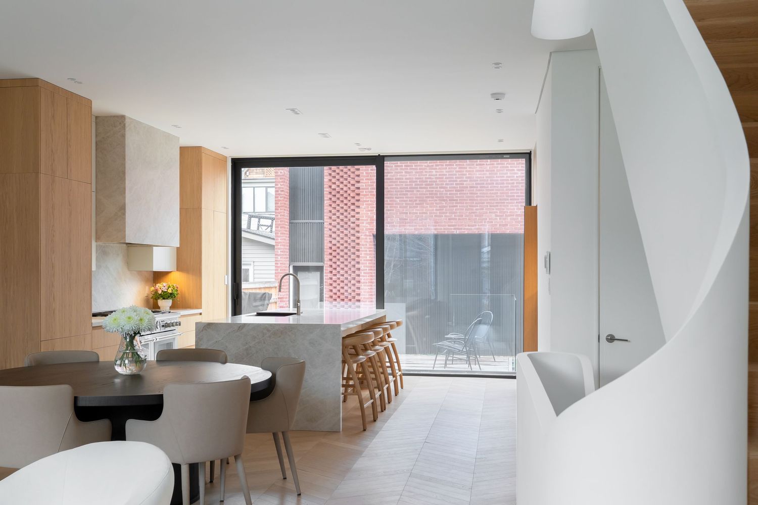 Contemporary open-plan kitchen and dining area with marble island, wooden cabinetry, and large window overlooking brick courtyard.