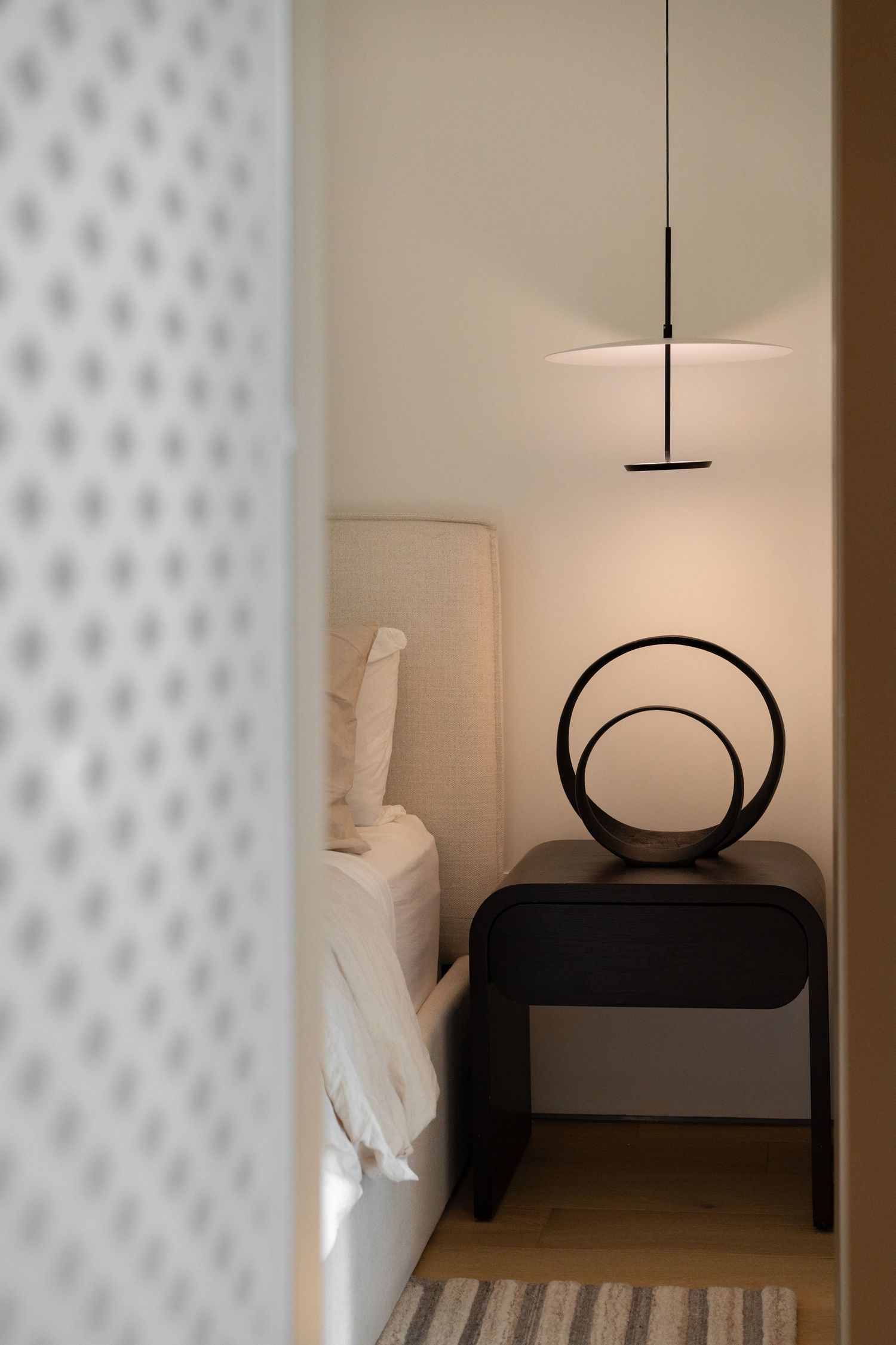 Modern bedroom with dark nightstand, circular lamp, and pendant light in a minimalist design.