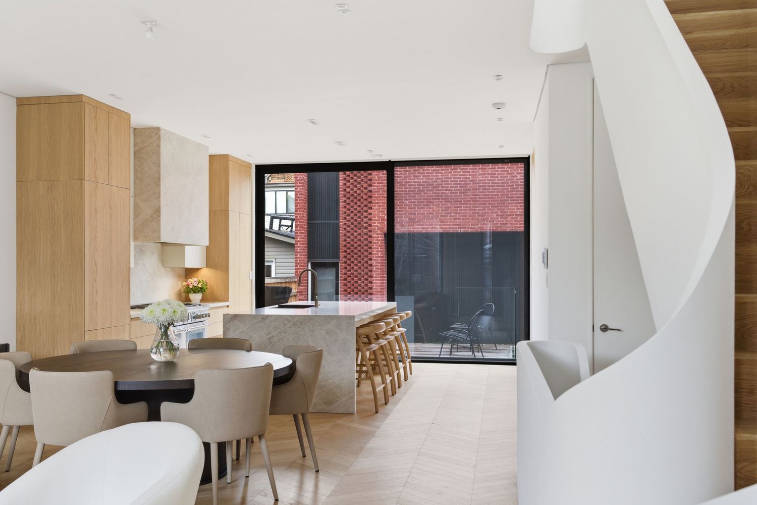 Modern open-plan kitchen and dining area with timber cabinetry, marble island, and large glass doors overlooking a brick courtyard.